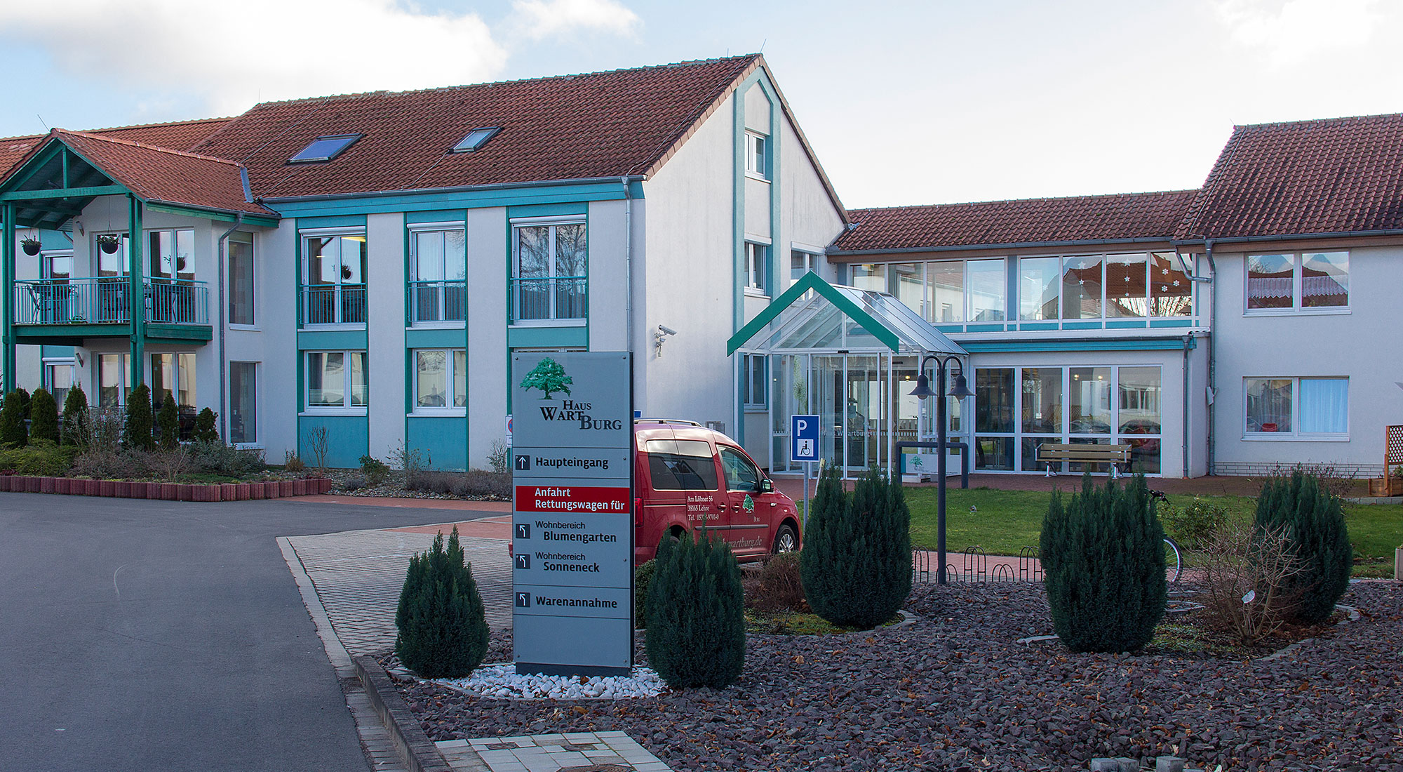 Außenansicht eines weißen Gebäudes mit hellblauen Verzierungen um die Fenster mit einem Wegweiser Schild im Vordergrund mit der Aufschrift Haus Wartburg