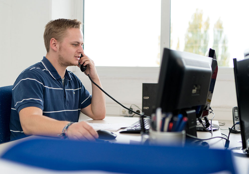 Kurzhaariger blonder Mann in dunkelblau-weiß gestreiftem Oberteil sitzt am Schreibtisch vor einem Desktop Computer und telefoniert mit einem schnurgebundenen Telefon in seiner linken Hand